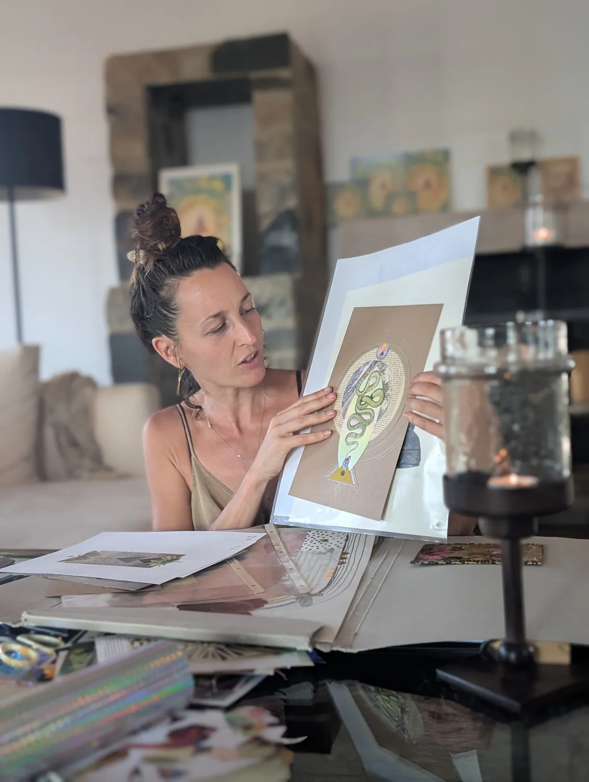 An artist holding up a finished collage, across a table spread with papers and materials